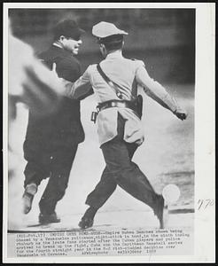Umpire Gets Bum-Rush -- Umpire Ruben Sanchez shown being chased by a Venezuelan policeman, night-stick in hand, in the ninth inning rhubarb as the irate fans started after the Cuban players and police arrived to break up the fight. Cuba won the Caribbean Baseball series for the fourth straight year in the 8-2 riot-studded decision over Venezuela at Caracas.
