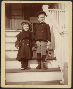 Two young children (girl and boy) standing on steps, boy is holding a pair of skates