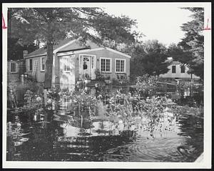 Beauty Amid Flood Damage is reflected here as flood waters of the Concord river spill into the attractive flower garden of Mrs. Helen Muskalski, 60, marooned in her home on Riverview (sic) avenue, Billerica. She is shown awaiting evacuation by fire department boats.