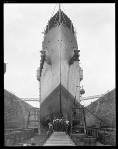 Repair crew poses in front of tall bow of the USS Raleigh in South Boston drydock