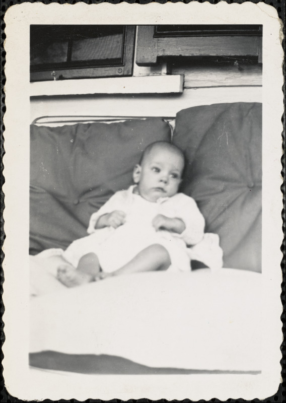 An infant dressed in white sits propped up against two large pillows