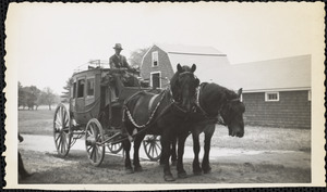 A man in a suit sits high in the driver's seat of a four-wheeled, dark-colored coach drawn by two large black horses