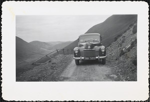 View of the front of a dark-colored car stopped on a dirt road in a mountainous landscape