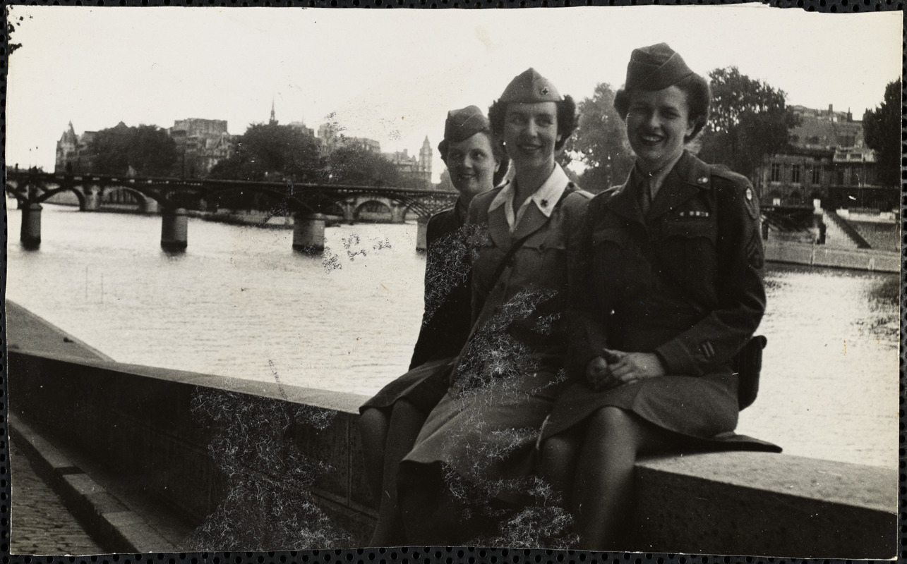 Three women, dressed in military-style uniforms, sit on a stone wall