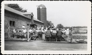 A gathering of men, women, and children sit at, or stand around, tables set up outside a shingled structure, most likely a farm building