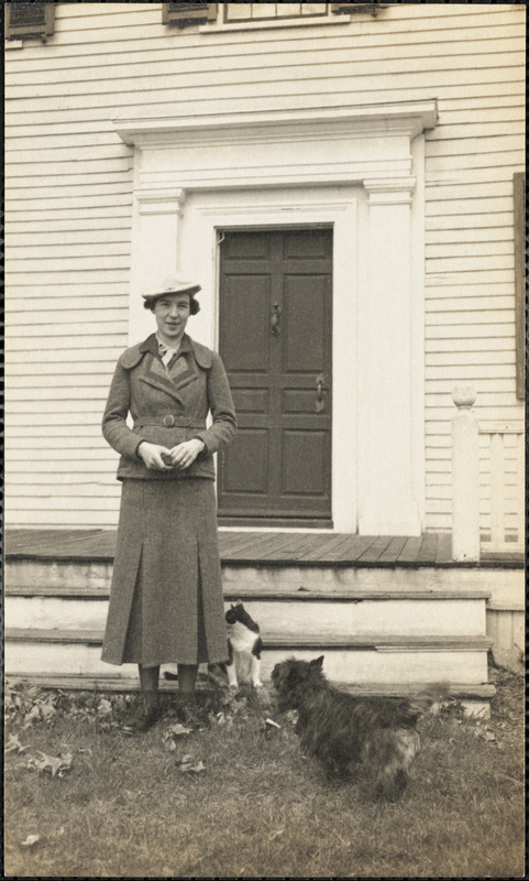 A woman stands in front of a porch with a cat and dog