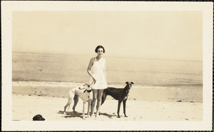Standing in the sand next to the ocean is a woman holding the leashes of two greyhounds