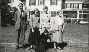 A man and three women stand with a German Shepard on the lawn in front of a light-colored house