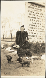 A woman stands in front of a large sign and holds the leashes of three dogs