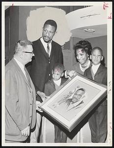 Celtics Great Bill Russell is presented a painting today at Hotel Lenox on the occasion of his being named Most Valuable Player in professional basketball for the third year in a row. Making award is Boston Garden basketball director Bill Mokray (left). With Bill are (left to right) son Jake, 3, wife Rose and son Buddha, 5.
