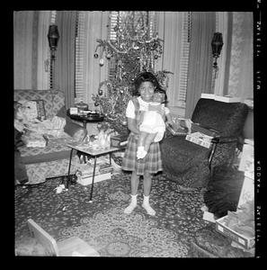 A girl stands holding a doll with a Christmas tree behind her