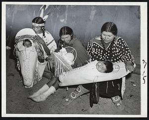 While Daddies Hunt-Three Shoshone Indian women with their babies await return of their husbands from antelope hunt at Lander, Wyo. The babies still ride the traditional boards on their mothers' backs. Left to right are Mrs. Ruby Tindall with 9-month-old Mary Ruby; Mrs. Tinnie Norman with 6-month-old Sylvester, and Mrs. Suzette Wagon with 9-month-old William. Soft white buckskin covers the papooses.