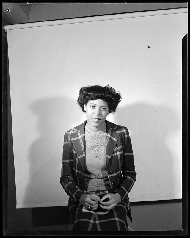 A woman sits on a stool, a white backdrop behind her