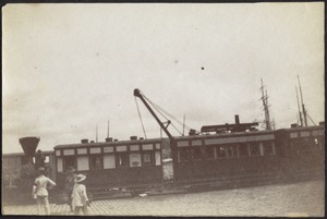 Train cars at harbor; people in foreground; ships masts in distance