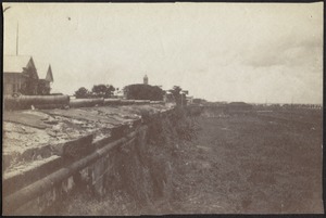 Side view of artillery cannons lined up along stone wall; buildings on right
