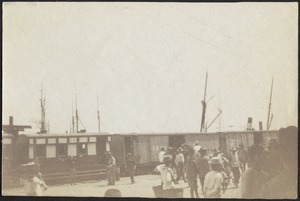 View of dock with train cars and sailing vessels; people walking about