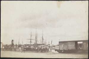 View of dock with train cars, sailing vessels and steamboat; people walking about