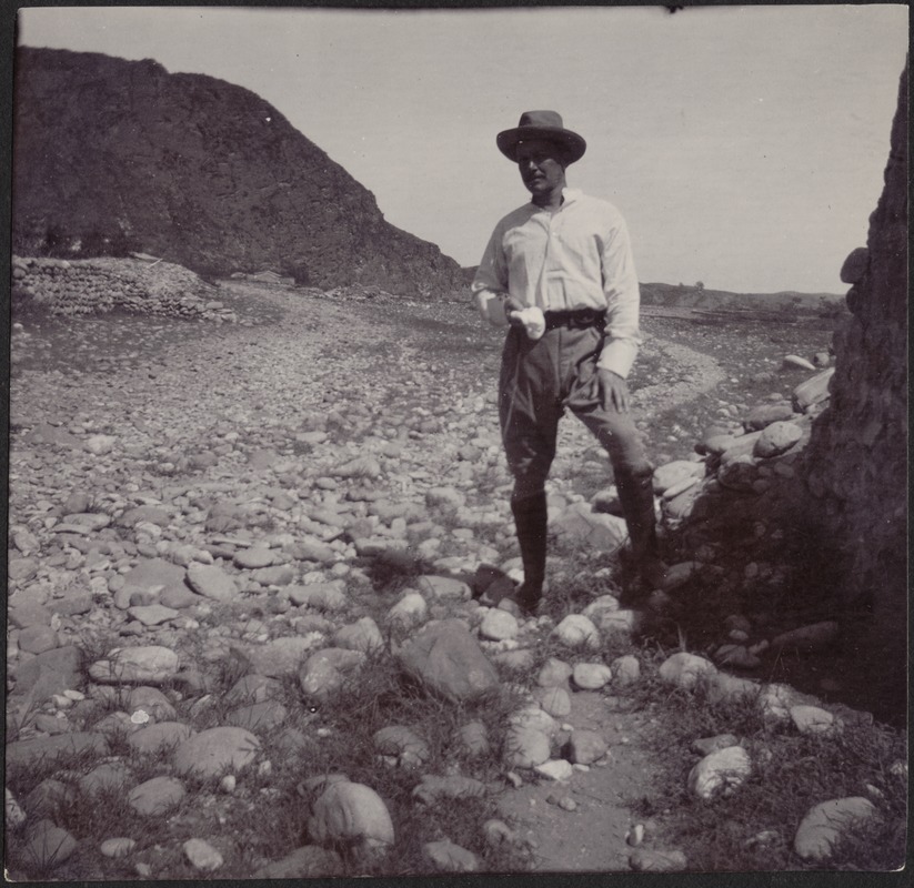 John Gardner Coolidge in wide-brimmed hat standing in a rocky, mountainous terrain; building in distance