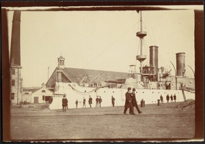 Battleship in dock; men in bowler hats walking about and observing