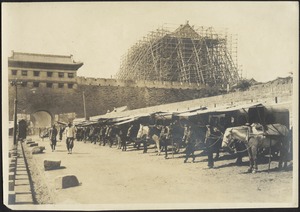 View of people walking down road outside of city gate with horse stables; building under construction behind crenelated wall