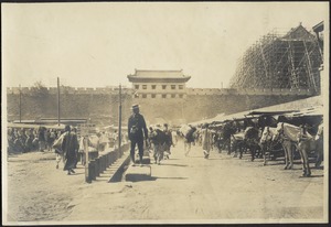 View of busy road outside of city gate/crenelated wall with horse stables; man in western dress standing in center; building under construction behind wall on right