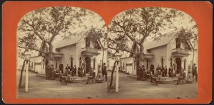 People on balcony and seated and standing outside of cottage
