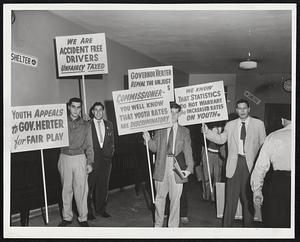Pickets, posters and conversation enlivened today’s hearing over automobile insurance rates. Here students display posters, which were ordered out of the hearing room.