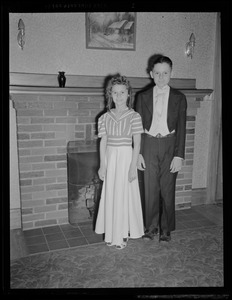 Unidentified girl and boy in formal wear standing in front of a fireplace