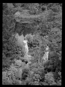 Two people standing on rocks, surrounded by trees, with water in the background