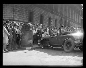 A crowd of people stand beside a damaged car, Boston Public Library in background