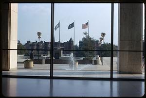 A view of three flags on flagpoles through glass, fountain in foreground