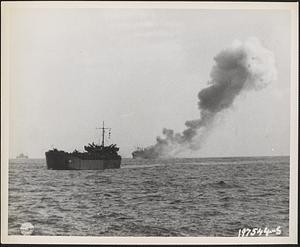 Struck by a Japanese torpedo plane, a liberty ship, in convoy off Leyte, is shown afire in the distance