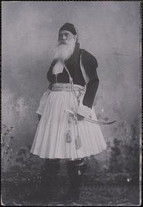 A bearded man stands wearing traditional Greek dress including a fustanella