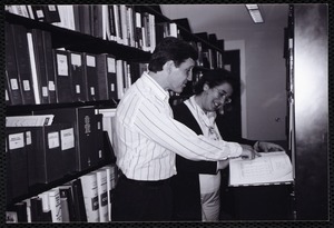Newton Free Library, Newton, MA. Communications & Programs Office. 2 staff checking reference book in stacks