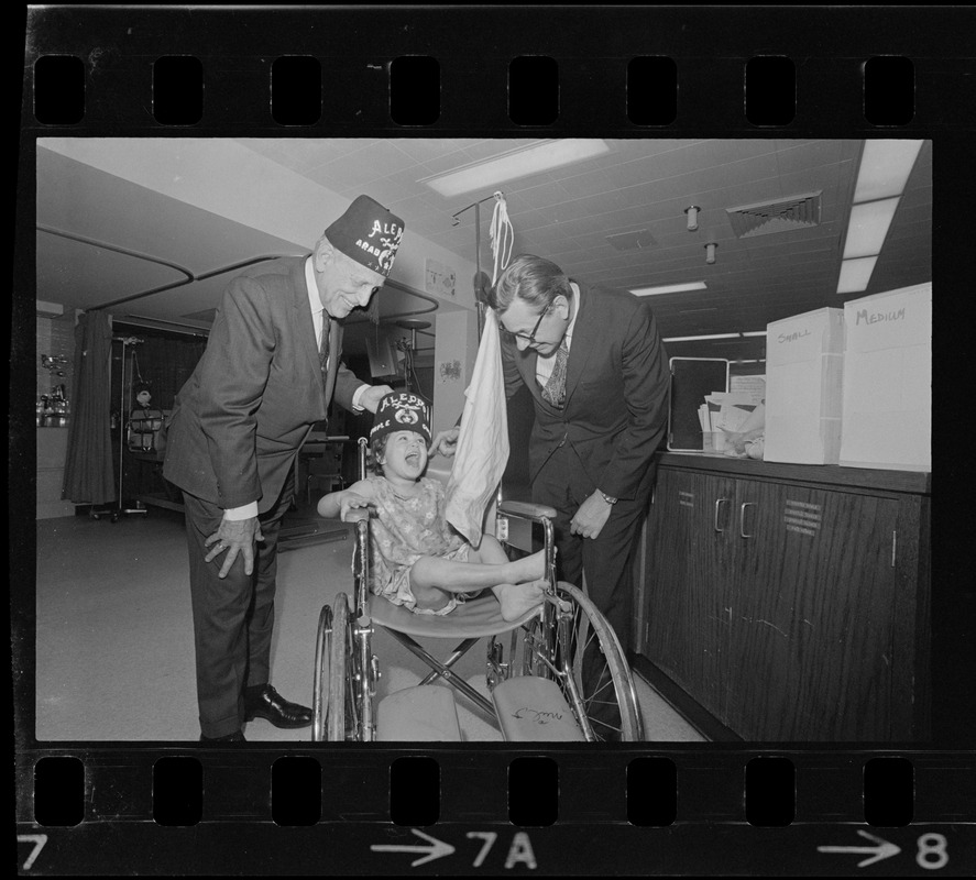 Judge Robert Gardiner Wilson, Jr., of Aleppo Temple, Boston, and Dr. John Burke, chief of staff of the Shriner Burn Hospital, places an Aleppo Temple fez on Veronica Diemont of Northampton, a patient at the hospital, during hospital day festivities