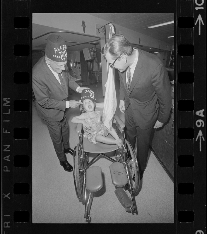 Judge Robert Gardiner Wilson, Jr., of Aleppo Temple, Boston, and Dr. John Burke, chief of staff of the Shriner Burn Hospital, places an Aleppo Temple fez on Veronica Diemont of Northampton, a patient at the hospital, during hospital day festivities