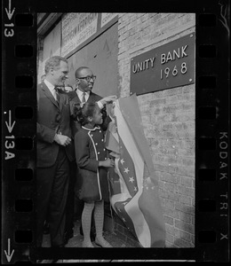 Renne Johnson, 8, the youngest stockholder of the Unity Bank and Trust Co., New England's first bi-racial bank, pulls off the flag which draped the bronze cornerstone plaque during unveiling ceremonies in Roxbury. Watching with delight is Mayor Kevin White, who was a guest speaker, and Donald Sneed, President and Chairman of the Board