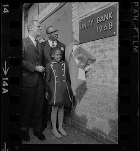 Renne Johnson, 8, the youngest stockholder of the Unity Bank and Trust Co., New England's first bi-racial bank, pulls off the flag which draped the bronze cornerstone plaque during unveiling ceremonies in Roxbury. Watching with delight is Mayor Kevin White, who was a guest speaker, and Donald Sneed, President and Chairman of the Board