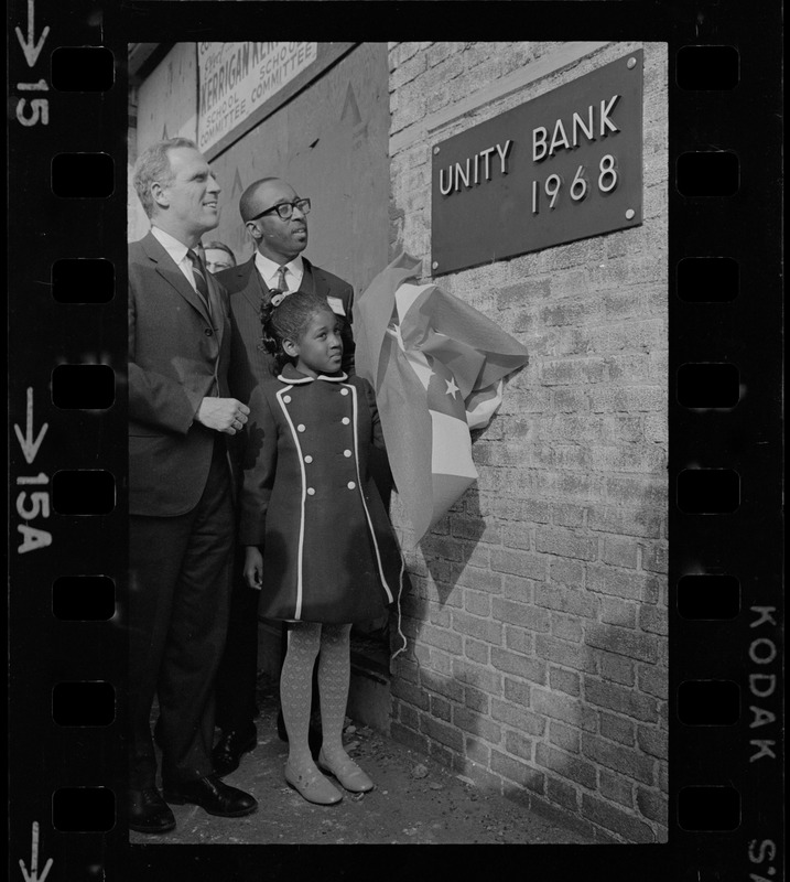 Renne Johnson, 8, the youngest stockholder of the Unity Bank and Trust Co., New England's first bi-racial bank, pulls off the flag which draped the bronze cornerstone plaque during unveiling ceremonies in Roxbury. Watching with delight is Mayor Kevin White, who was a guest speaker, and Donald Sneed, President and Chairman of the Board