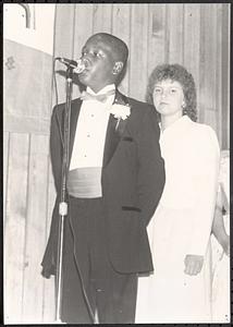 Richard "Mokey" Harding wearing a tuxedo and standing at microphone, with a young women behind him