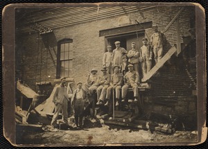 Group of laborers, some holding tools, posed on a staircase outside of a building