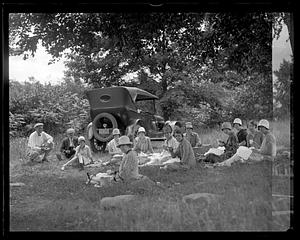 A group of people picnicking, an automobile in the background