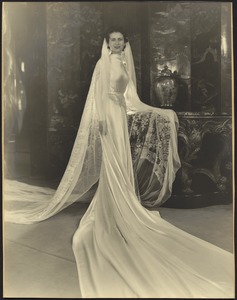 Woman in wedding dress with long draped train and veil; oriental furnishings in background