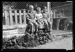 Three boys sit in front of a chain-link fence