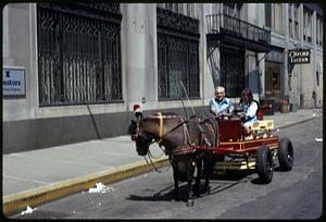 Two people on a horse drawn cart going down city street