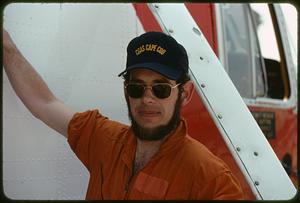 Man wearing "CGAS Cape Cod" hat standing next to helicopter