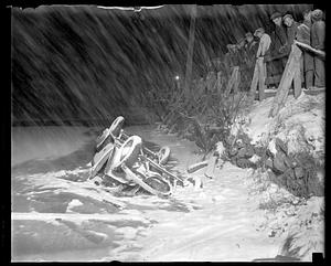 People looking over a fence at an overturned automobile in icy water during a snowstorm