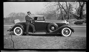 A man in suit stands in front of a car with holding a cigarette