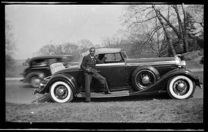 A man in a suit holding his hat stands in front of a car