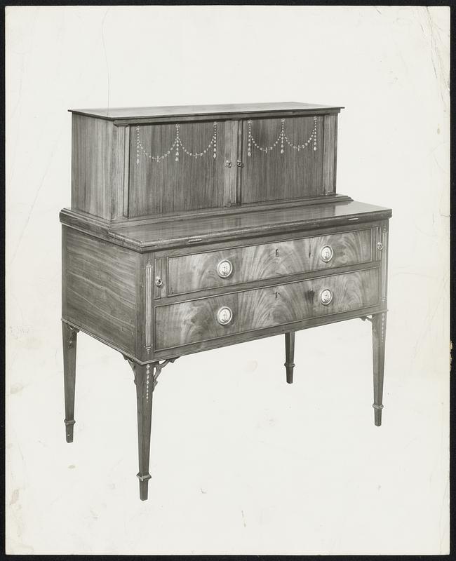 Tambour desk by John Seymour & Son ca. 1800, Boston. Height 3'5". One of the choice examples of New England furniture of late 18th century. Constructed of San Domingan mahogany with satinwood inlay, the inlaid pilaster forms and sliding tambour shutters are characteristic of Seymour's work. The inlaid festoons on the reeded shutters are details rarely found. The four ring handles frame circular plaques of Bilston enamel. Two drawers and three pigeon holes behind each tambour shutter.